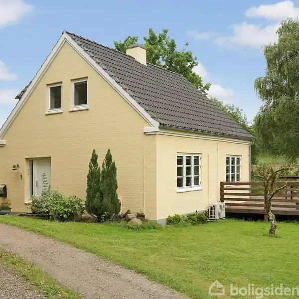 Hus i beige mursten står stille på græsplæne med lille træterrasse. En grusvej fører forbi huset, omgivet af grønne træer og buske under en delvist skyet, blå himmel.