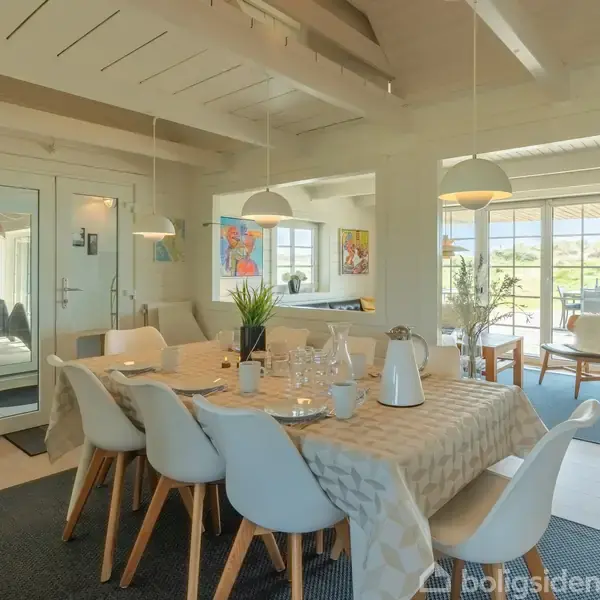 Spisebord med hvid dug dækket med glas og kande. Rummet er lyst og moderne med hængende lamper og store vinduer, der viser en terrasse med udsigt til landskab.