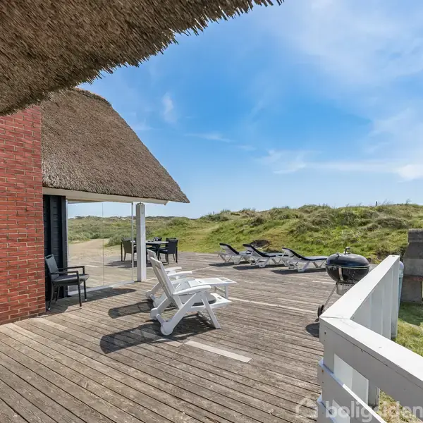 Stråtagshus set fra terrasse, hvor to hvide liggestole står. Grill og ekstra stole på træterrassen, omgivet af græs og klitter under en blå himmel.