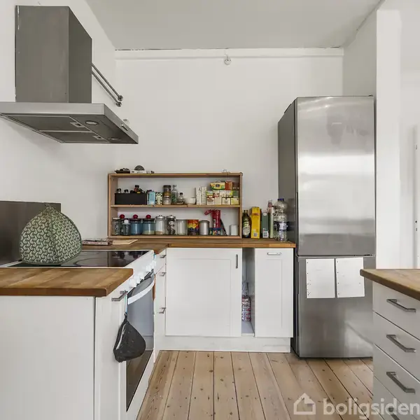 Et moderne køkken med hvide skabe og træbordplader, indeholdende en stor stålkøleskab, ovn og en hylde med krydderier og madvarer. Rummets gulv er af lyst træ.