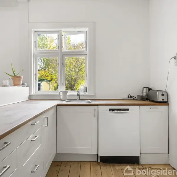 Hvidt køkken med træbordplade. En plante og en brødrister står på bordet. Et vindue med naturligt lys viser grønne træer udenfor. Trægulv og opvaskemaskine er synlige.