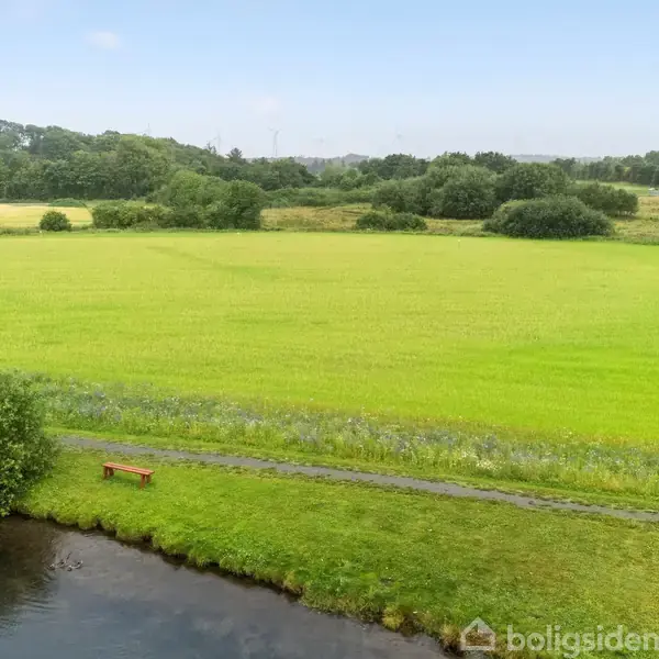 Bænk står stille ved en flodbred, omgivet af frodige grønne marker under en klar himmel. I baggrunden ses træer og buske, der skaber en rolig, naturlig atmosfære.