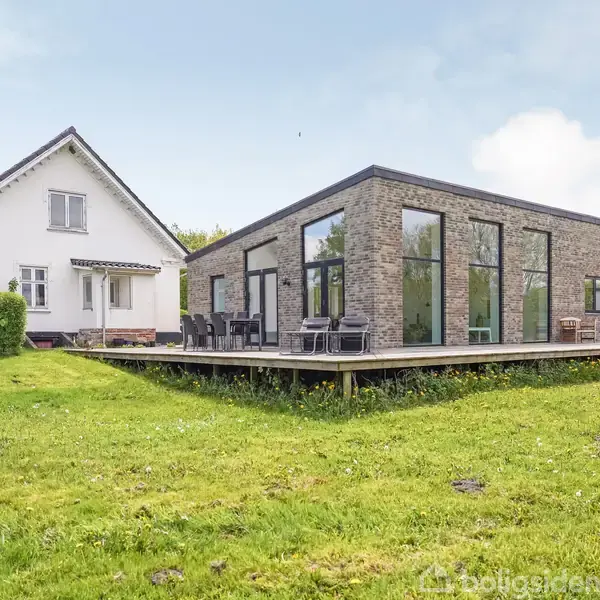 To huse står i en have med græsplæne. Det ene hus er moderne med store vinduer og terrasse, mens det andet er traditionelt med skråt tag.