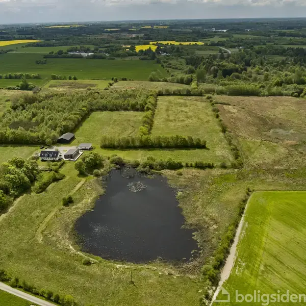 En bygning ligger blandt træer ved siden af en stor dam. Det hele er omgivet af marker og landbrugsjord.
