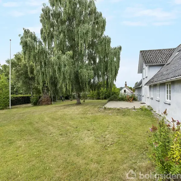 Et hvidt hus med mørkt tag står side om side med en stor grøn græsplæne. Store træer omgiver ejendommen under en klar blå himmel.