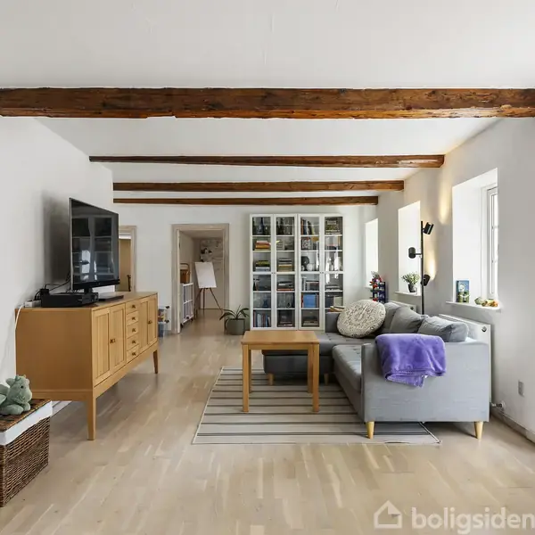 En stue med gråt sofahjørne og TV på skænk. Træbjælker i loftet. Sofaen har en lilla plaid, og der står en gulvlampe ved siden af. Lyse trægulve og store vinduer.