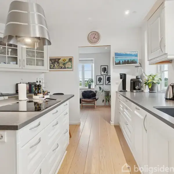 Et lyst køkken med hvide skabe og sort bordplade. Bordet har køkkenudstyr, og der er vask, elkoger og kaffemaskine ved vinduet. Billeder hænger på de hvide vægge.