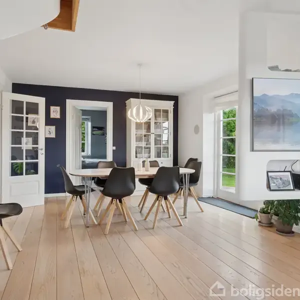 Et moderne køkken med hvide skabe og sorte bordplader. En kaffemaskine og en elkedel står ved vasken. Trægulvet fører til en lys stue. Vægkunst dekorerer væggene.