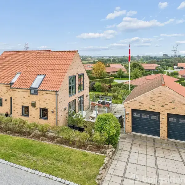 Et murstenshus med rødt tegl, to etager og flere ovenlysvinduer. Ved siden er der en garage med to døre. Græsplæne og terrasse omgiver huset, med en flagstang i baggrunden.