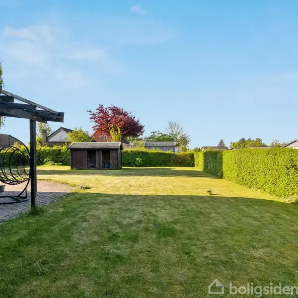 En velplejet have med græsplæne omgivet af grønne hække. Et rødt murstenshus har en terrasse med en sort pergola og moderne gyngestole. Vejret er solrigt og klart.