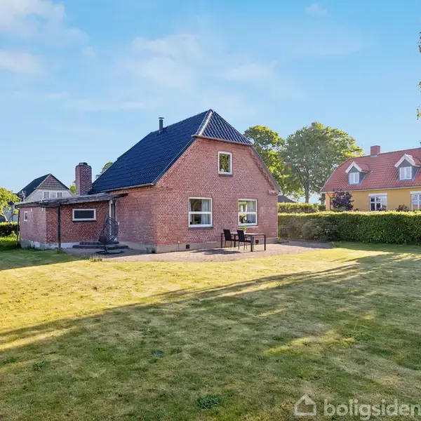 Murstenshus står stille i en stor, velplejet have med klippet græs og omkranset af hække. En flisebelagt terrasse ved husets bagindgang har et bord og stole.