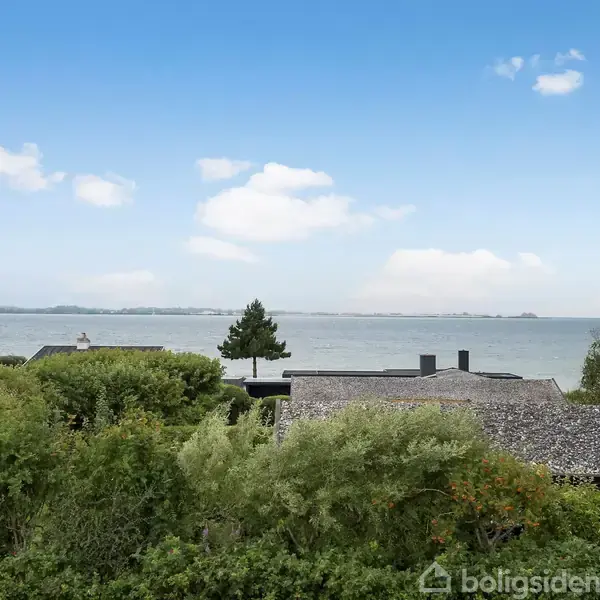 Husene ligger stille blandt frodig bevoksning ved kysten med udsigt til havet under en klar blå himmel med enkelte skyer.