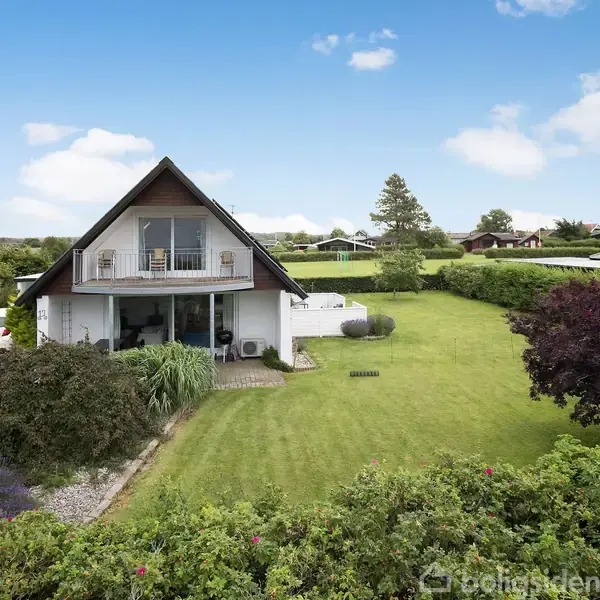 Et hvidt, to-etagers hus med spidst tag står i en velplejet have. En bil holder i indkørslen til venstre, omkranset af træer og buske under en blå himmel.