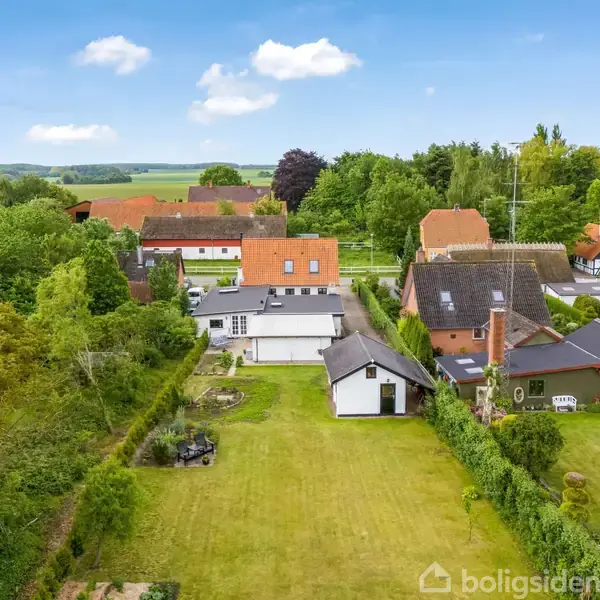 En hvid bygning med sort tag står i en frodig have. Omkring den er der flere huse med orange tage, træer og marker under en blå himmel med skyer.