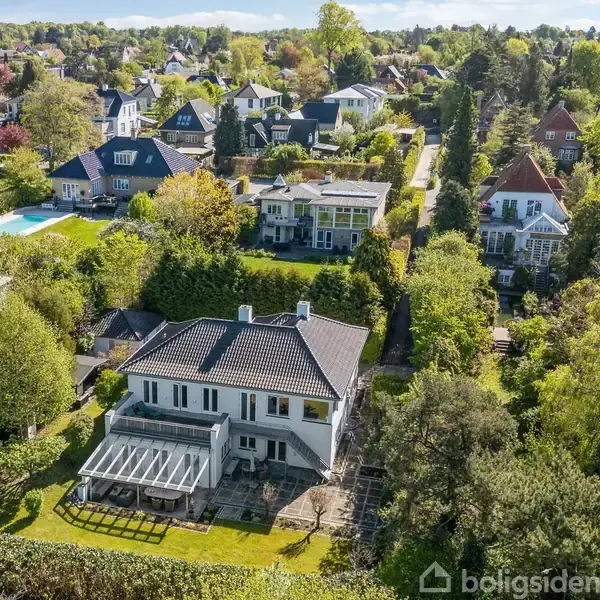 Hus i forgrunden med stor have og terrasse, omgivet af frodige træer. Baggrunden viser en række lignende huse i et grønt boligområde.