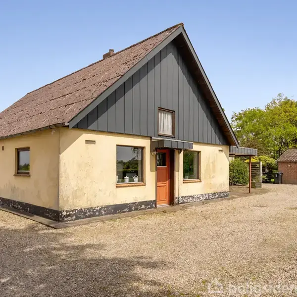 Et gult murstenshus med et mørkt tag og store vinduer står stille på en grusplads, omgivet af grønne træer og en udhusbygning i baggrunden.