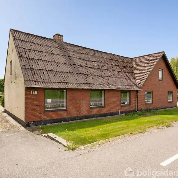 Et rødt murstenshus med skråt tag står ved en vej. Huset har tre store vinduer og en græsplæne foran med træer i baggrunden. Skilt med tallet 77 på væggen.