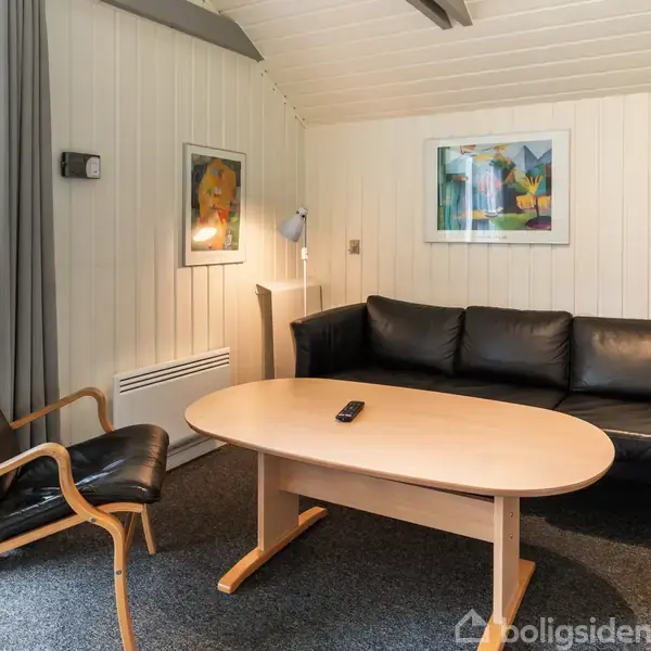 Sort lædersofa og lænestol står i et lyst stuehjørne med trævægge. Et ovalt træbord har en fjernbetjening. Væggene er dekoreret med farverige billeder.