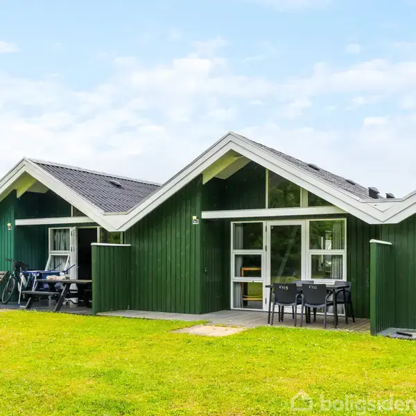Grønt sommerhus med skråtag ligger på en grøn græsplæne. Der står havemøbler foran huset, og det er omgivet af en lys, skyet himmel.