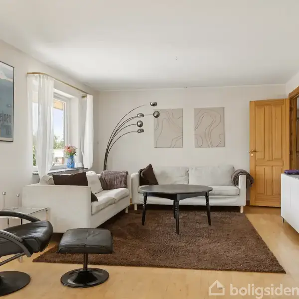 Stue med en sort lænestol, hvid sofa og rektangulært sofabord på et brunt tæppe. Store vinduer tilfører lys. En TV-skænk står til højre; kunst dekorerer væggene.