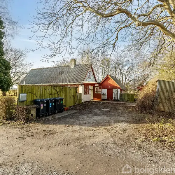 Et lille hus med rødt og hvidt træværk står stille omgivet af nøgne træer og en grusvej. Fire skraldespande står op ad en grøn trævæg.