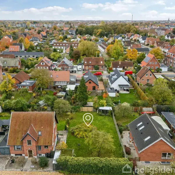 Et parcelhusområde med røde tegltage og grønne haver set fra oven. Huse omgiver en central have markeret med en nåleikon. Omgivende huse og træer skaber en idyllisk byudsigt.