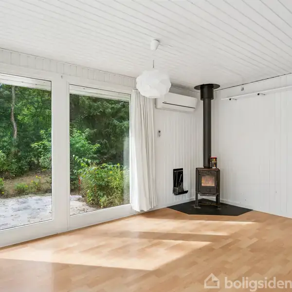 En lys stue med trægulv har en brændeovn ved væggen. Store vinduer slipper udsigt til grøn have ind. Hængende hvid lampe i loftet og en airconditionenhed på væggen.