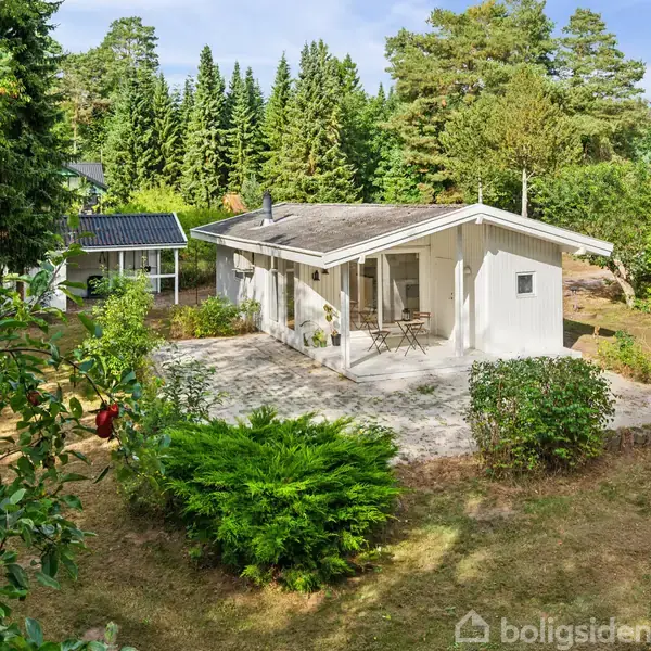 Et hvidt hus står stille i en solrig have omkranset af grønne træer og buske. En flisebelagt terrasse med havemøbler er foran huset, omgivet af velplejet græs.