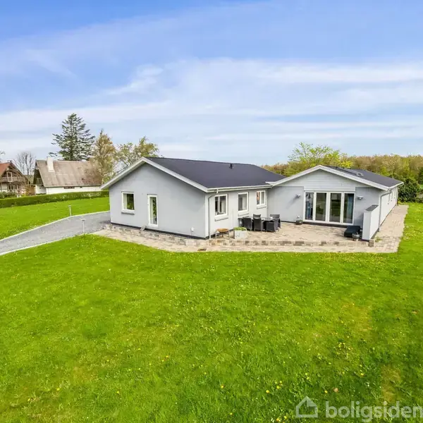 Huset står stille på en frodig grøn græsplæne med en flisebelagt terrasse og havemøbler. Omkring ligger et åbent grønt område med få træer og en klar blå himmel.