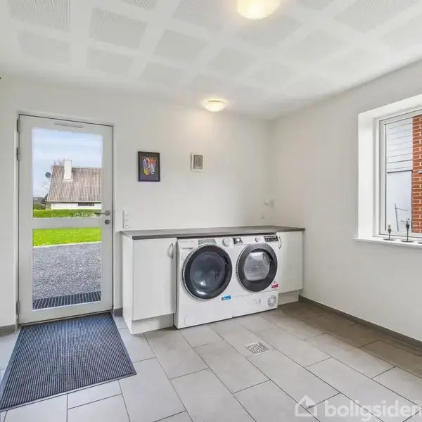 Vaskemaskine og tørretumbler står side om side på en hvid bordplade i et lyst bryggers med flisegulv og udsigt til grøn have gennem et vindue ved siden af hoveddøren.