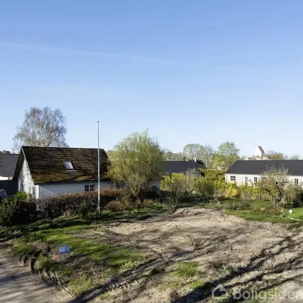 Et hus med mørkt tag står ved en grusvej, omgivet af grønne træer og buske; i baggrunden ses flere huse og blå himmel.