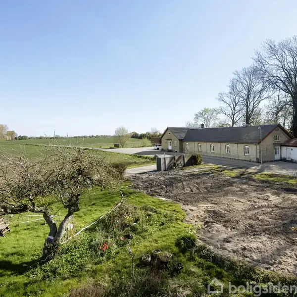 En lang, lys boligbygning står stille ved siden af en åben mark og skovområde. Foran bygningen er der en bar jordområde, mens en græsplæne med træ og havemøbler ses til venstre.