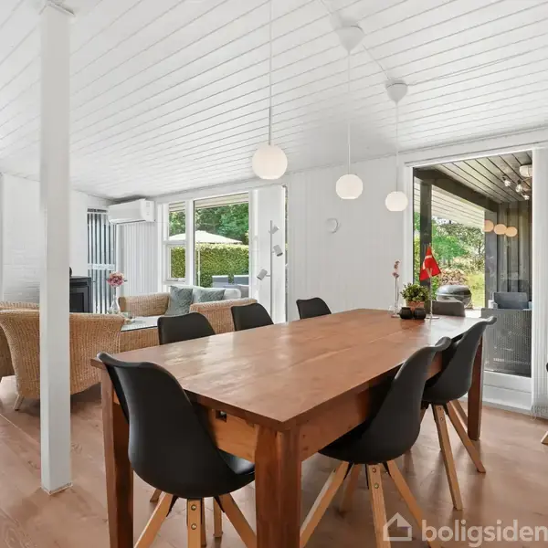 Spisebord i træ med sorte stole omkring står i en moderne stue. Store vinduer og glasdøre giver udsigt til frodig have. Lyse lofter og gulve skaber en lys atmosfære.