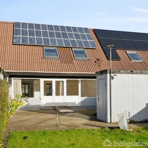 Rækkehus med solpaneler på taget, nogle åbne vinduer; have med grøn græsplæne og flisebelagt terrasse. En skurbygning er placeret til højre.