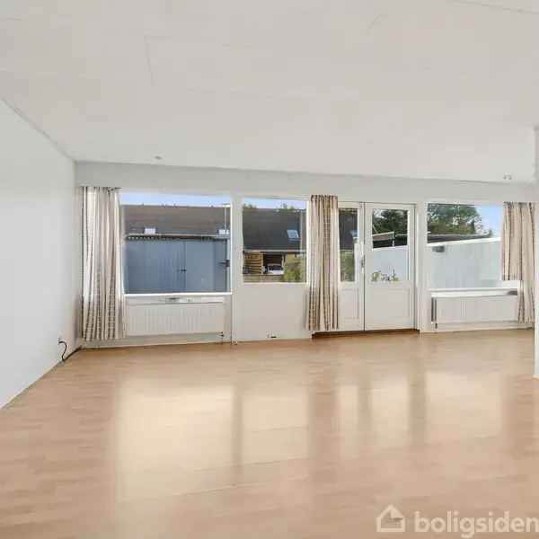 Tomt rum med trægulv, store vinduer og en glasdør med udsigt til udendørs områder. Lyse vægge og gardiner skaber en lys og rummelig atmosfære.