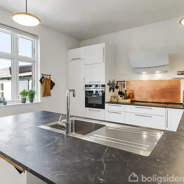 Køkken med sort bordplade og integreret vask, omgivet af hvide skabe og ovn. Kobberfarvet backsplash tilføjer farve. Store vinduer giver naturligt lys, omgivet af pæne dekorationer.