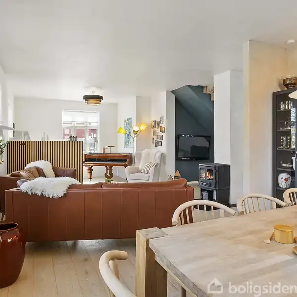 En stue med en brun sofa står ved et sofabord. I baggrunden ses en pejs, spisebord og stole. Rummet er lyst og har moderne møblering.