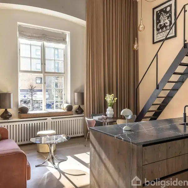 Loftseng i stue med trappe, moderne indretning. Under, en lyserød sofa ved stort vindue. Køkkenø med sort bordplade, to pendellamper, maleri på væggen.