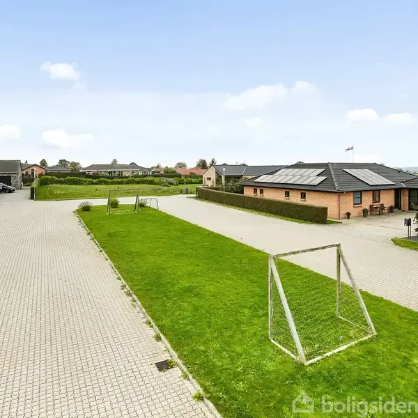 Et åbent græsareal med et fodboldmål står på en villavej. Omgivet af flere huse med flade tage og solpaneler samt stenbrobelagt vej under en blå himmel.