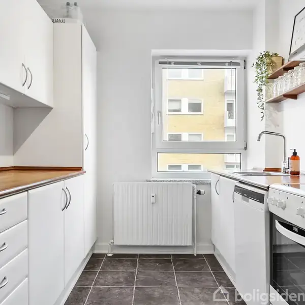 Et lyst køkken med hvide skabe og skuffer. Et komfur og opvaskemaskine er integreret. En enkeltstående vask ved et vindue fylder det lille rum.