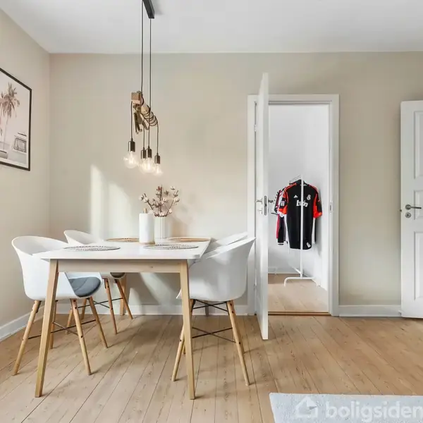 Bord med fire stole står i en lys spisestue. Bordet er dækket med magasiner og en vase. I baggrunden ses åbne døre ind til et værelse med tøj på bøjler.