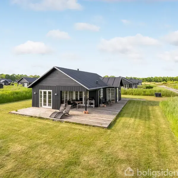 Sort sommerhus står på stor græsplæne med træterrasse og liggestole. Omkringliggende huse set i baggrunden under en skyet himmel.