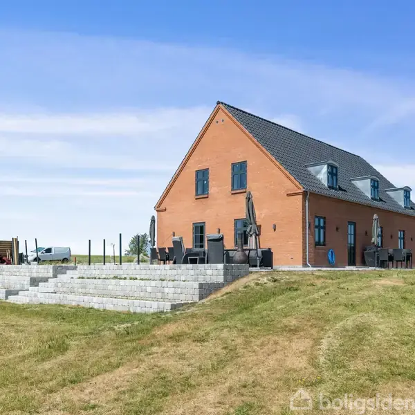 En stor, moderne murstensvilla står på en hævet græsplæne med terrasse og havemøbler i forgrunden. Baggrunden er klar blå himmel og grøn mark.