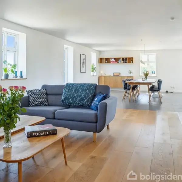 Sofa med puder står i en moderne stue med trægulv. Et sofabord har en blomsterbuket og bogen "FORMEL 1." I baggrunden ses et spisebord og reoler.