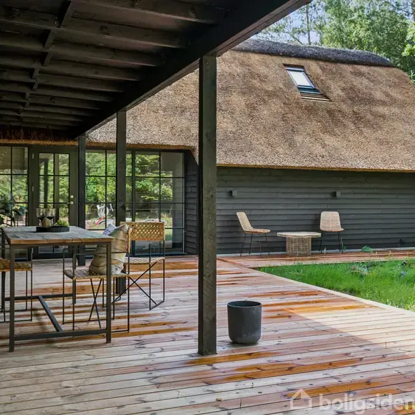 Træterrasse med spisebord og stole, placeret foran et hus med stråtag. Omgivet af grønne planter og en skov. Pejs og pynteting kan ses på bordet.