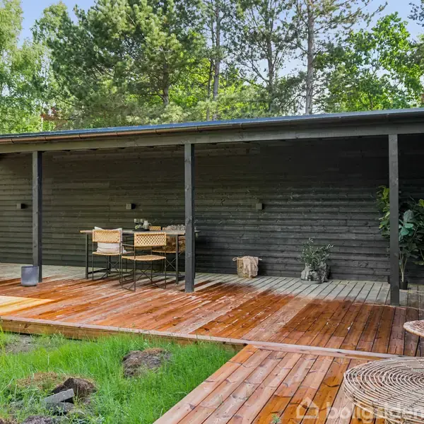 En træterrasse med møbler foran et sortmalet sommerhus. Udendørs spisebord og stole står under et halvtag. Omgivet af frodig skov med træer i baggrunden.