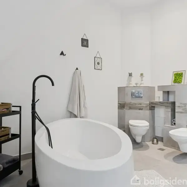 Et fritstående badekar med en sort armatur står i et lyst badeværelse. Til højre er der et ophængt toilet og en bidet ved siden af en dekoreret hylde.