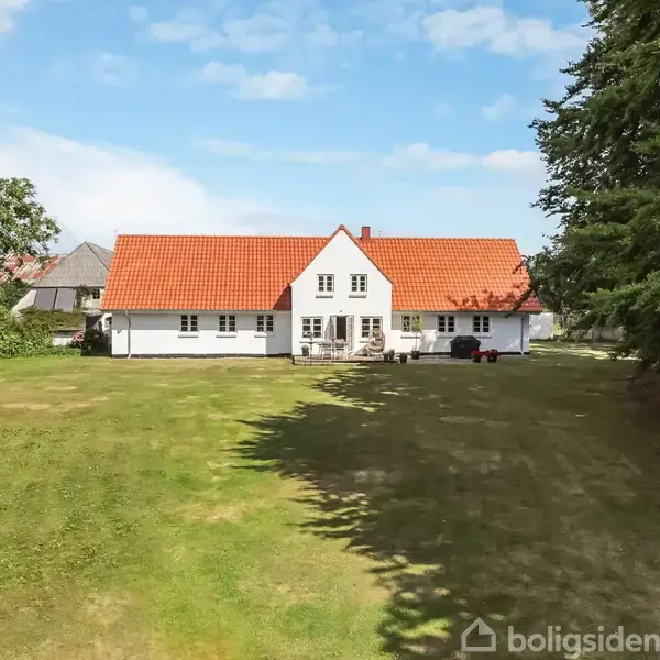 Et hvidt hus med rødt tegltag står på en stor græsplæne omgivet af træer og buske under en blå himmel med skyer.