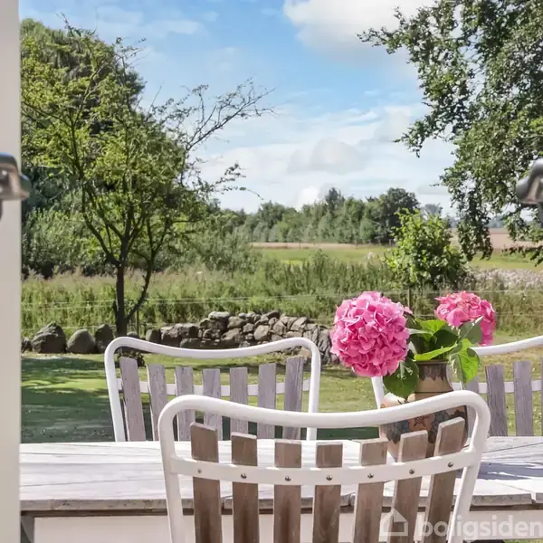 Et udendørs bord med pink blomster i vase står omgivet af lyse træstole. Udsigten består af en frodig have med træer og en stengærde i baggrunden under en blå himmel.