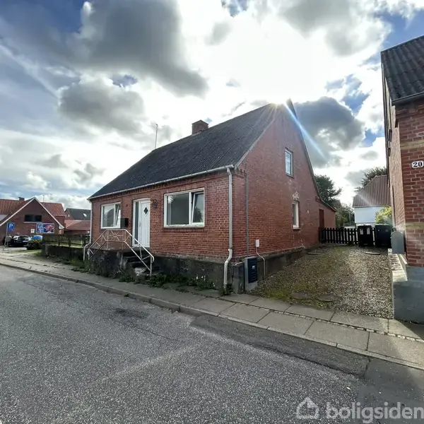 Rød murstenshus med sort tag står stille langs en villavej med fortov og andre lignende huse. Sollys bryder gennem skyerne, hvilket skaber en lys atmosfære.